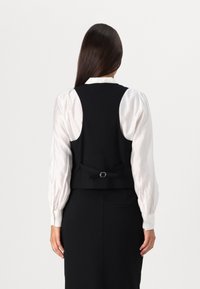 Black tailored vest with a cinched back and silver hardware detail, worn over a white blouse with puffed sleeves. Smooth fabric texture.