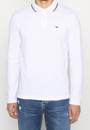 Homme portant un polo blanc à manches longues avec un col rayé noir et un petit logo sur la poitrine, associé à un jean bleu déchiré.