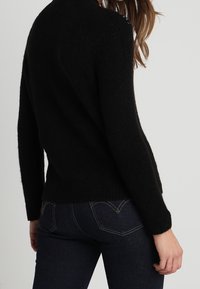 Schwarzer gestrickter Pullover mit strukturierter Oberfläche und hohem Kragen. Verfügt über blaue Stickdetails an den Schultern und einen leicht gekürzten Saum.