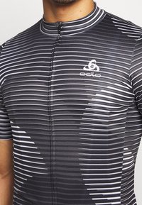 Cyklistický dres s krátkým rukávem vyrobený z elastické tkaniny, s černo-bílým pruhovaným vzorem, vysokým límcem a předním zipem.