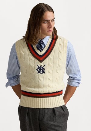 Man with shoulder-length hair wearing a striped blue shirt, navy tie, and cream cable-knit sweater vest with red and navy stripes.