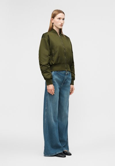 Γυναίκα που στέκεται φορώντας πράσινο μπουφάν bomber, φαρδιά μπλε τζιν και μαύρα παπούτσια, μπροστά σε απλό ανοιχτό γκρι φόντο.