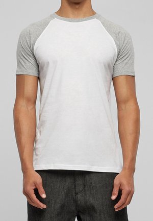 Mand iført hvid kortærmet raglan-T-shirt med lysegrå ærmer og mørkegrå bukser, stående mod en ensfarvet grå baggrund.