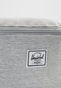 Gros plan d’un sac en tissu texturé gris clair avec une fermeture éclair visible et un patch avec le logo de la marque Herschel Supply Co.