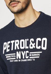 Muž v tmavě modrém tričku s bílým textem "PETROL&CO NYC Brooklyn U.S. Bez bariér Rychle a s kontrolou od '73"