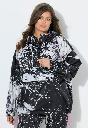 Kvinde iført sort og hvid sprøjtmønster vindjakke med lyserøde snore, stående mod en hvid baggrund.