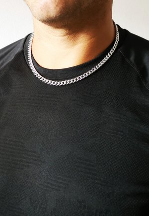 Sudraba ķēdes kaklarota uz personas, kas valkā melnu teksturētu kreklu, ar bieziem saitēm un gludu virsmu.