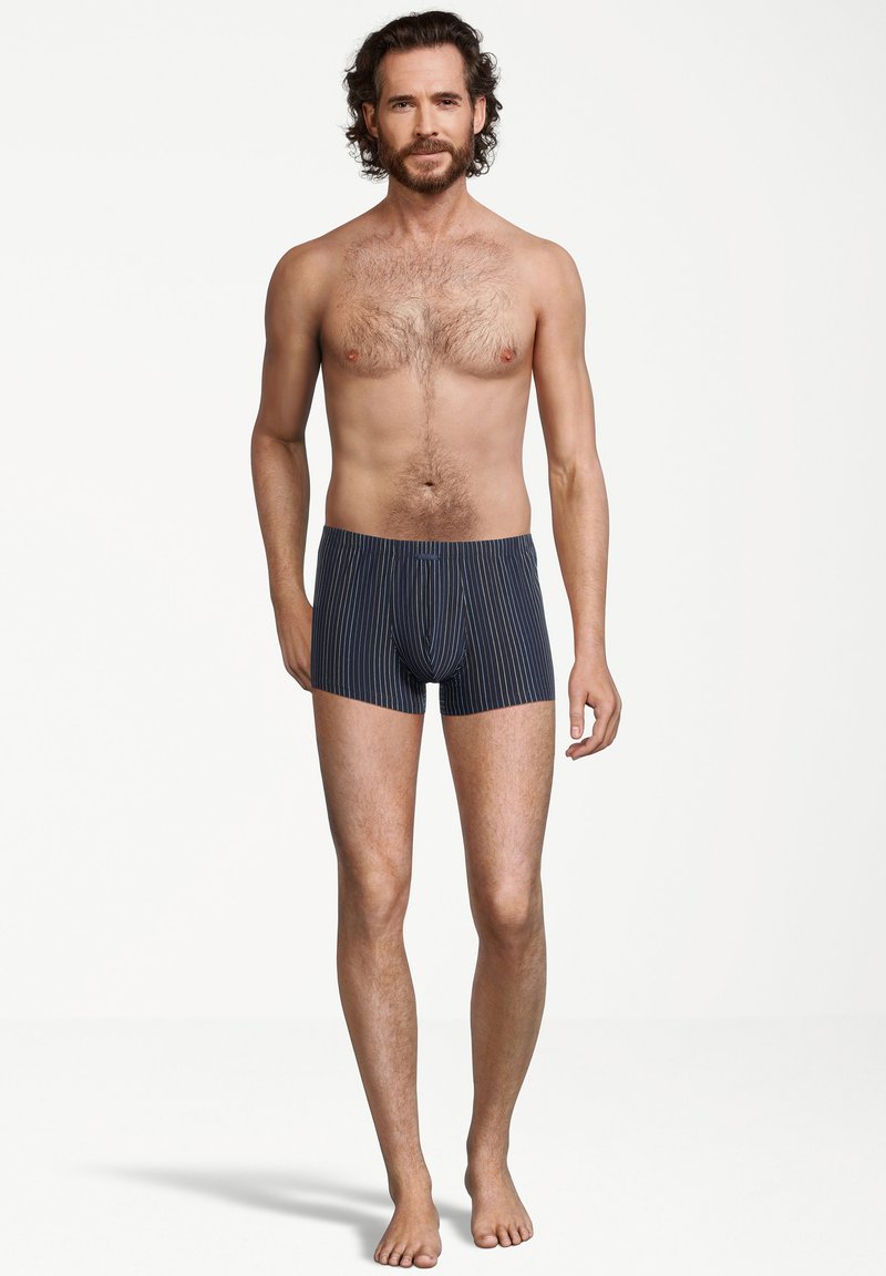 Bearded man met krullend haar staat blootsvoets in marineblauwe boxershorts met dunne witte verticale strepen tegen een effen witte achtergrond.