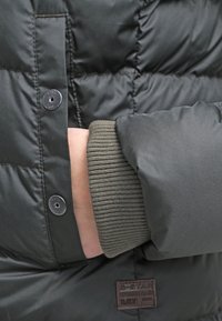 Ręka w kieszeni czarnej pikowanej kurtki z prążkowanym mankietem i widocznymi zatrzaskami, pokazująca część brązowej tkaninowej metki przy dolnym brzegu.