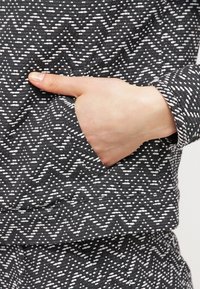 Černobílá látka s vzory cikcak a puntíků. Ruka se objevuje v boční kapse, čímž zdůrazňuje funkčnost oděvu.