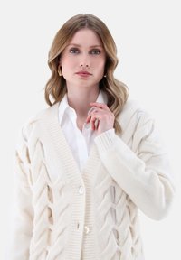 Strickcardigan in Creme mit strukturiertem Zopfmuster, V-Ausschnitt und Druckknopfverschluss, mit gerippten Abschlüssen; über einem weißen Hemd getragen.