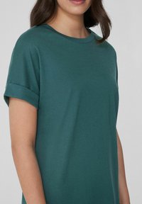 Tyrkysový top s krátkým rukávem a kulatým výstřihem, vyrobený z měkké bavlny. Má ohrnuté manžety a volný střih, bez viditelných vzorů.