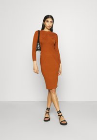 Haljina od rebrastog pletenog materijala u boji hrđe, dugih rukava, uskog kroja i dijagonalnim detaljima. Uparena s crnim sandalama na remen i malom torbicom.