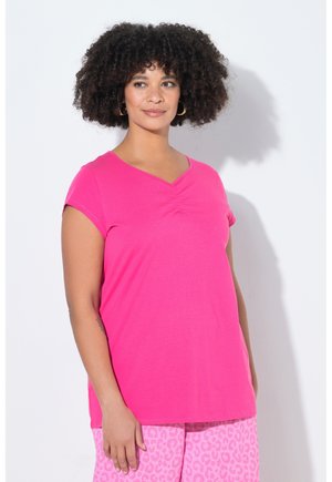 Frau mit lockigem Haar, die goldene Creolen, ein leuchtend pinkes Kurzarmoberteil und hellrosa Leopardenmuster-Hosen trägt, steht vor einer weißen Wand.