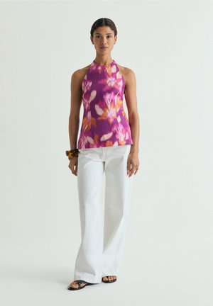 Mujer que lleva un top halter sin mangas floral en púrpura y rosa, pantalones blancos de pata ancha, sandalias negras y pulseras marrones, de pie.