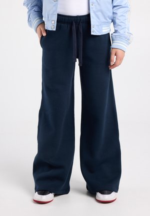 Persoon draagt marineblauwe wijde sweatpants, witte sneakers met rode zolen en een lichtblauw jack met gestreepte manchetten.