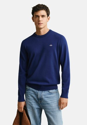 Junger Mann trägt einen dunkelblauen Pullover mit einem kleinen Emblem auf der Brust, hellblaue Jeans und hält eine braune Jacke in einer Hand.