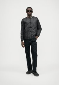 Giacca nera trapuntata con colletto alto e chiusure a toggle, abbinata a pantaloni scuri e scarpe slip-on, indossata con occhiali da sole.