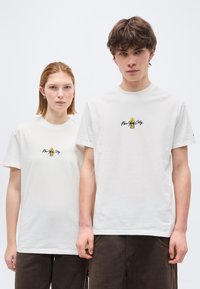 Δύο νέοι ενήλικες φορούν λευκά μπλουζάκια με το γράφημα "New York City" και σκοτεινό παντελόνι, στέκονται δίπλα δίπλα μπροστά σε ένα απλό φόντο.