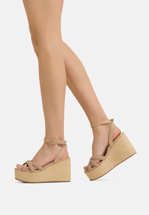 Beine tragen beige gewebte Plateau-Wedgesandalen mit Knöchelriemen vor einem einfarbigen weißen Hintergrund.