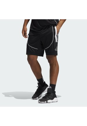 Mężczyzna w czarnych krótkich spodenkach sportowych z białym wykończeniem, czarnych skarpetkach Adidas i czarnych butach sportowych z marmurkowymi podeszwami, stojący z jedną ręką w kieszeni.