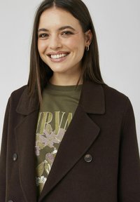 Manteau croisé marron foncé avec un col cranté, en matière texturée, porté sur un t-shirt vert à motifs graphiques avec des éléments floraux.