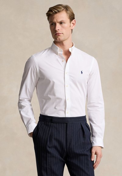 Homme portant une chemise blanche boutonnée et un pantalon bleu marine à fines rayures, debout les mains dans les poches sur un fond neutre.