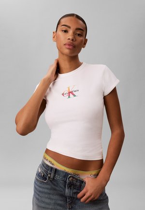 Žena v bielom krátkom tričku Calvin Klein a sivých džínsoch, ukazujúca žltý pás s logom Calvin Klein, pózujúca na sivom pozadí.