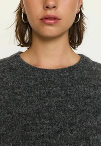 Tamsiai pilkas vilnonis megztinis su tekstūruotu paviršiumi, turintis plačią iškirptę ir minkštą, pūkuotą paviršių. Papuoštas minimalistiniais žiediniais auskarais.