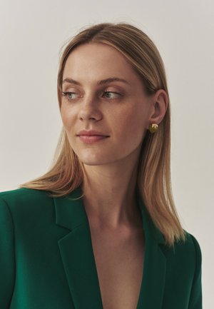 Blazer verde con scollatura a V profonda, caratterizzato da un design strutturato. Orecchini a perno color oro con finitura testurizzata decorano l'orecchio del modello.