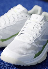 Coppia di scarpe sportive bianche Adidas con strisce grigio chiaro e dettagli verdi posate su una superficie blu strutturata.