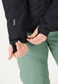 Schwarze wasserdichte Jacke mit verstellbaren Bündchen, mit glatter Stoffstruktur und sicherem Klettverschluss, getragen über grünen Hosen.