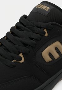 Primer plano de una zapatilla negra con gruesos cordones negros y un logotipo "etnies" en color marrón en la lengüeta y el panel lateral.