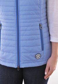 Blau gesteppte Weste mit einem gepunkteten Muster, Reißverschlusstaschen und einem runden Logo-Patch. Mit einem hohen Kragen und kontrastierenden blauen Reißverschlüssen.