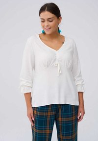 Witte, lichtgewicht blouse met een V-hals, geplooide voorkant en een trekkoord. Lange mouwen zijn gegatherd bij de manchetten. Gecombineerd met groen geruite broek.