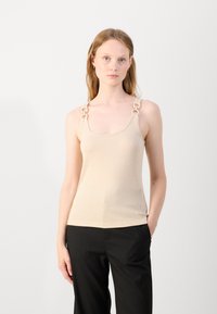 Beige tank top with ribbed texture and wide shoulder straps adorned with circular hardware. Fitted silhouette, paired with black trousers.