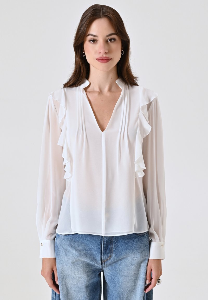 Blusa bianca a maniche lunghe con dettagli arricciati lungo il colletto. Tessuto leggero, design a V e polsini con bottoni. Abbinata a jeans in denim blu.