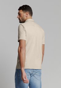 Beige polo-shirt met korte mouwen en een kraag, van structuurstof met een rechte zoom. Gecombineerd met lichtblauwe spijkerbroek, getoond vanuit de achterkant.