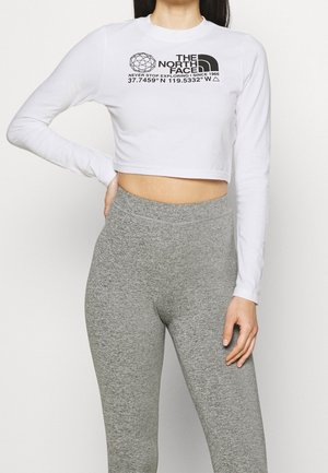 Nainen, joka pukeutunut valkoiseen lyhennettyyn pitkähihnaiseen toppiin, jossa on The North Face -logo, ja harmaisiin korkeavyötäröisiin legginsseihin, seisoo yhdellä kädellä lanteilla.