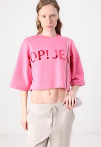 Pink cropped sweatshirt with "POP! JEAN" in reflective letters, wide sleeves, paired with light gray drawstring pants and a small handbag.