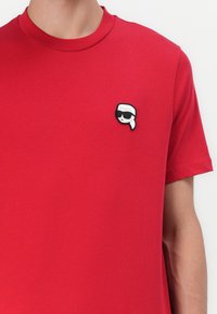 Raudona medvilninė marškinė su apvalia iškirpte ir trumpomis rankovėmis. Krūtinėje yra juodai baltas piešinys, vaizduojantis figūrą akinių nuo saulės.