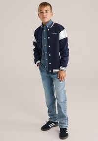 Jūras spēku sporta žakete ar baltām akcentiem, valkāta pāri gaiši zilai krekliņam ar pogām. Kombinēta ar nolietotām gaiši zilām džinsām un melniem sporta apaviem.