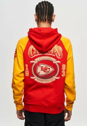 Haut à capuche rouge et jaune avec une poche kangourou, comportant un design graphique au dos et des manches de couleur contrastée. Tissu doux et texturé.