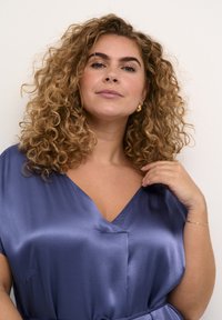 Rochie din satin de un albastru închis, cu mâneci scurte și decolteu în V. Prezintă o talie strânsă și buzunare laterale. Modelul are păr creț și cercei din aur.