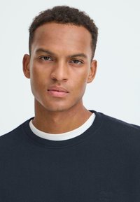 Marineblauer Sweatshirt mit rundem Ausschnitt und strukturiertem Stoff. Hat einen weißen Innenkragen und ein kleines Logo-Detail auf der Brust.