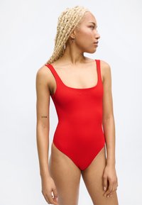 Noor naine blondide palmikutega, kandes punast üheosalist ujumistrikood, vaadates kõrvale, vasakul käsivarrel on tätoveering "DIVINE".