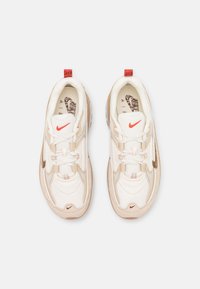 Sneakers z białą siateczką i beżowymi panelami z zamszu, z czerwonym akcentem na pięcie i charakterystycznym złotym logo w kształcie "swoosh".