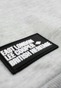 Tissu gris à surface texturée présentant un patch en caoutchouc noir avec un texte blanc en relief : "EAST LONDON LEE COOPER BRITISH HERITAGE".