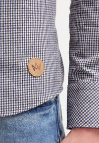 Dettaglio di una camicia a quadri marrone e blu con una toppa in pelle che presenta un logo, un colletto con polsini e uno sfondo in denim.