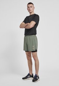 Sort t-shirt med korte ærmer, olivengrønne atletiske shorts med en sideslids og lysegrå chevronmønster, parret med sorte og blå sneakers.